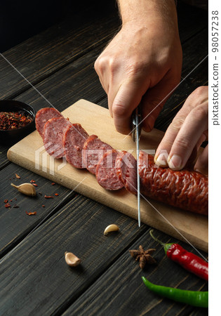 Cutting sausage on wooden cutting board at domestic kitchen by hands chef for a sandwich. Cutting sausage on wooden cutting board at domestic kitchen by hands chef for a sandwich. 98057238