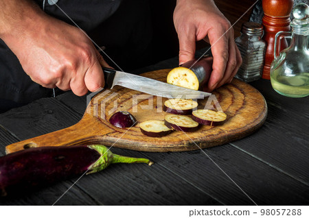 Professional chef is cutting an eggplant in the restaurant kitchen. Close-up of the hands of the cook during work. Delicious breakfast or dinner 98057288