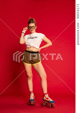 Stylish coach. Portrait of fashionable young girl in t-shirt and shorts rollerblading over red background. Sport, fashion, happy emotions and active lifestyle concept 98058171