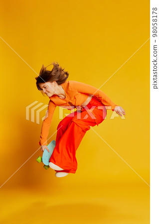Expressive girl in orange croptop and trousers with skateboard jumping over colorful yellow background. Youth, fashion, actvie lifestyle, happy emotions and active lifestyle concept 98058178