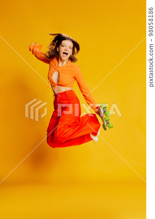 Expressive girl in orange croptop and trousers with skateboard jumping over colorful yellow background. Youth, fashion, actvie lifestyle, happy emotions and active lifestyle concept 98058186