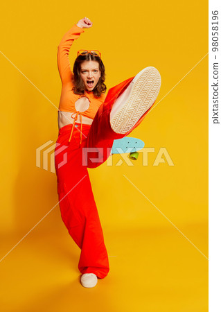 Expressive girl in orange croptop and trousers with skateboard jumping over colorful yellow background. Youth, fashion, actvie lifestyle, happy emotions and active lifestyle concept Expressive girl in orange croptop and trousers with skateboard jumping over colorful yellow background. Youth, fashion, actvie lifestyle, happy emotions and active lifestyle concept 98058196