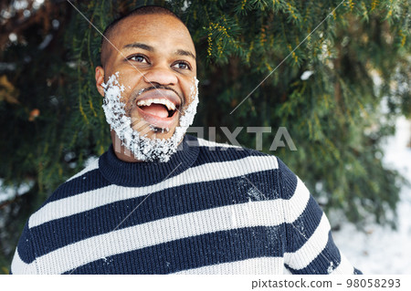 Cheerful funny African man with snow in beard enjoying winter weather, wearing striped sweater. Street portrait in park with snow. Lifestyle, city life concept. Close up, copy space 98058293