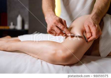 Man doing guasha massage of leg using IASTM tool for treatment of myofascial restriction 98059375