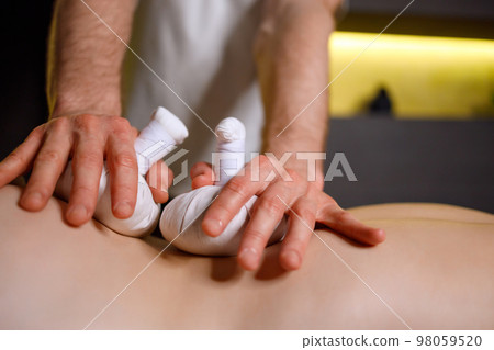 Close-up view of male masseur's hands putting herbal bags on female back during Thai massage 98059520