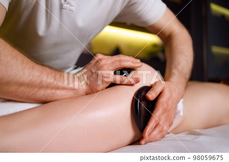 Man doing a massage session with warm stones on the buttocks and thighs of a female client of a spa resort Man doing a massage session with warm stones on the buttocks and thighs of a female client of a spa resort 98059675