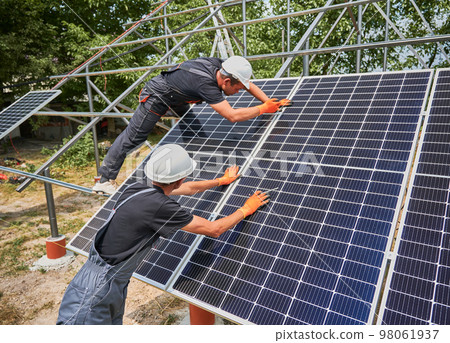Workers installing photovoltaic solar panel system outdoors. Men engineers placing solar module on metal rails, wearing construction helmets and work gloves. Renewable and ecological energy. 98061937