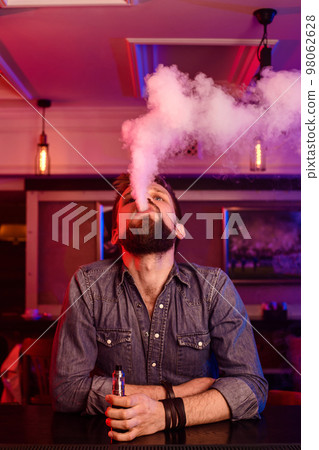 Vape. Vaping man in a cloud of vapor. Photo is taken in a vape bar. Vape. Vaping man in a cloud of vapor. Photo is taken in a vape bar. 98062628