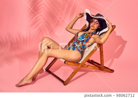 Summer lifestyle fashion portrait of young stunning woman on a lounge chair, pink background, studio shot. Wearing stylish sunglasses, bikini. Summer lifestyle fashion portrait of young stunning woman on a lounge chair, pink background, studio shot. Wearing stylish sunglasses, bikini. 98062712