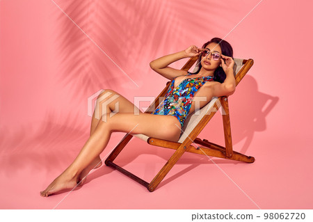 Summer lifestyle fashion portrait of young stunning woman on a lounge chair, pink background, studio shot. Wearing stylish sunglasses, bikini. Summer lifestyle fashion portrait of young stunning woman on a lounge chair, pink background, studio shot. Wearing stylish sunglasses, bikini. 98062720