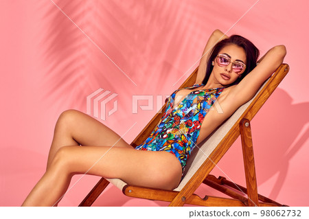 Summer lifestyle fashion portrait of young stunning woman on a lounge chair, pink background, studio shot. Wearing stylish sunglasses, bikini. 98062732
