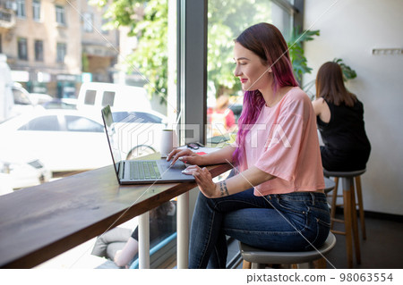Side view of young businesswoman sitting at table in coffee shop. On table cup of coffee and laptop. In background white wall and window. 98063554