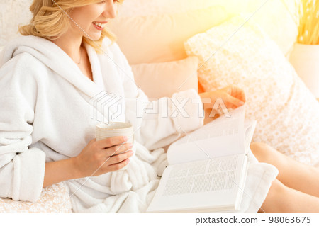 Young woman in white terry robe is drinking coffee and reading magazine or book in bedroom. Young woman in white terry robe is drinking coffee and reading magazine or book in bedroom. 98063675