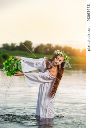 Beautiful black haired girl in white vintage dress and wreath of flowers standing in water of lake. Sun flare. 98063688