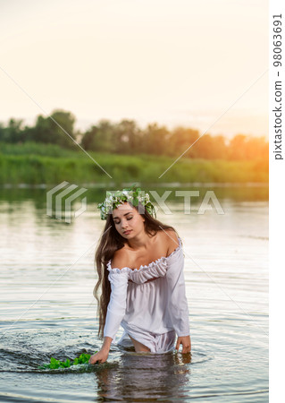 Beautiful black haired girl in white vintage dress and wreath of flowers standing in water of lake. Sun flare. 98063691