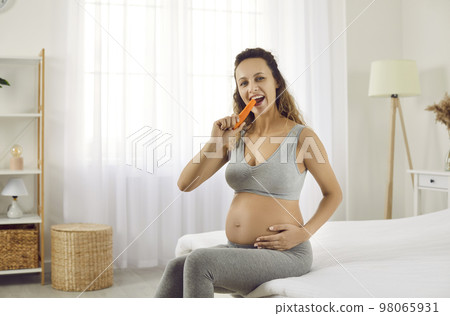 Happy young pregnant woman sitting in the bedroom and eating a fresh organic carrot 98065931