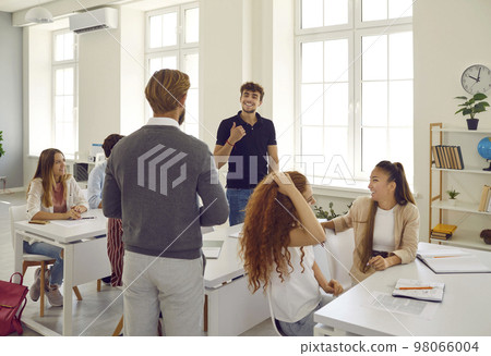 Bearded teacher communicates with student through desk against background of large bright window in modern classroom. Bearded teacher communicates with student through desk against background of large bright window in modern classroom. 98066004
