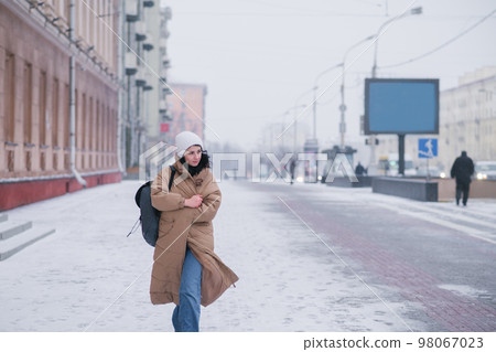 A girl in a long coat in cold weather with the wind is walking around the city 98067023