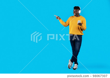Black Man Wearing Headphones Holding Smartphone In Hand And Pointing Aside 98067307