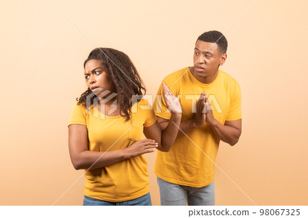 Black husband trying to talk and reconcile with offended wife, standing over yellow studio background 98067325