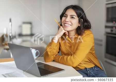 Dreamy Young Arab Female Sitting At Desk With Laptop In Kitchen Dreamy Young Arab Female Sitting At Desk With Laptop In Kitchen 98067335