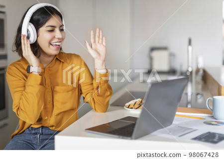 Young Arab Female Wearing Headphones Making Video Call On Laptop In Kitchen 98067428