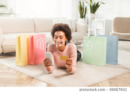 Cheerful young mixed race lady shopaholic with phone, credit card checking financial account lies on floor 98067602