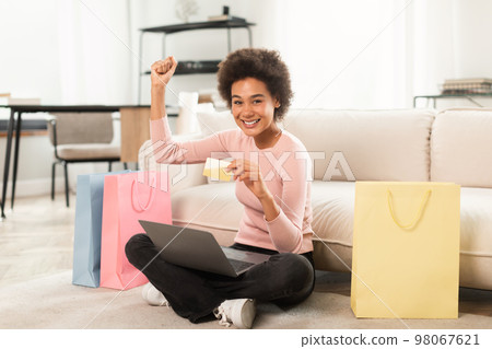 Satisfied young mixed race female shopaholic sit on floor with laptop, credit card, raising hand up celebrating success 98067621