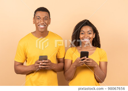 Portrait of happy black couple using cellphones and smiling at camera, standing on yellow studio background 98067656