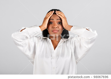 Confused shocked millennial black businesswoman in white blouse holding her hands behind head 98067696
