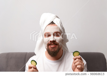 Portrait of funny happy man in white tshirt applying cucumber slices over clay mask on his face. Self care morning spa procedure 98068613