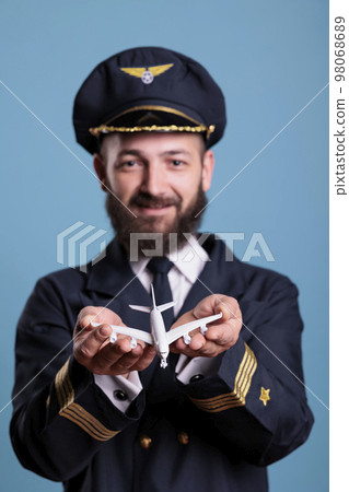 Portrait of airplane aviator in uniform holding plane toy in palms front view, pilot playing with commercial passenger jet model . Professional aviation academy aviator on blue background 98068689