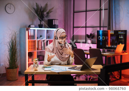 Young woman in headset having video call with partners at evening time, working process at office. Young woman in headset having video call with partners at evening time, working process at office. 98068760