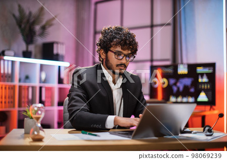 Confident bearded man in formal suit and glasses sitting at desk with modern laptop Confident bearded man in formal suit and glasses sitting at desk with modern laptop 98069239