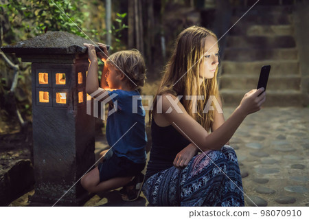 Mom looks into her smartphone, the son looks into the luminous cozy window of the house. The boy lacks Attention, care, love and home comfort 98070910