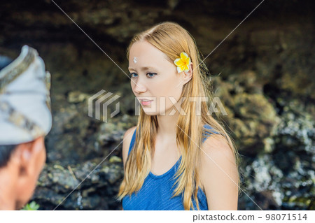 Woman is a tourist in Bali. The ritual of ablution with holy water and on her forehead prikleeli rice and put the flower ear Woman is a tourist in Bali. The ritual of ablution with holy water and on her forehead prikleeli rice and put the flower ear 98071514