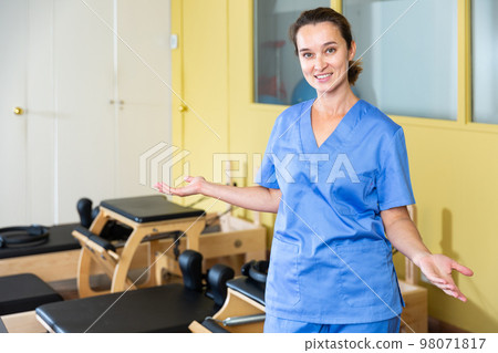 Smiling female physiotherapist inviting to rehab center with pilates equipment Smiling female physiotherapist inviting to rehab center with pilates equipment 98071817