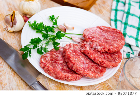 Fresh beef burgers with a leaf of parsley on a white plate placed on the wooden tabletop with garlic and kitchenware Fresh beef burgers with a leaf of parsley on a white plate placed on the wooden tabletop with garlic and kitchenware 98071877