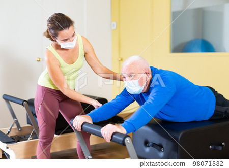 Senior man in protective mask exercising on Pilates reformer with female trainer 98071882