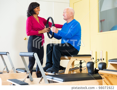 Hispanic woman fitness trainer controlling elderly man exercising with Magic Circle 98071922