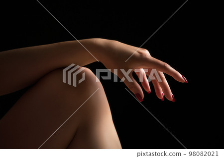 Woman's hand lies relaxed on bare knee on her leg. Crop closeup shot of relaxed female fingers with red nails on black background. Selective and soft focus on human hand. 98082021