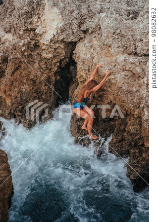A young beautiful tanned woman with long hair in blue bikini sitting at rocks near blue green clear ocean sea water. Woman relax on sunset at sea, enjoy holidays and weekend vacation in summer time. A young beautiful tanned woman with long hair in blue bikini sitting at rocks near blue green clear ocean sea water. Woman relax on sunset at sea, enjoy holidays and weekend vacation in summer time. 98082632