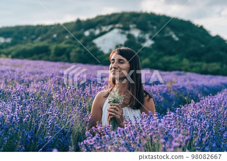 Close up portrait of happy young brunette woman in white dress on blooming fragrant lavender fields with endless rows. Warm sunset light. Bushes of lavender purple aromatic flowers on lavender fields. Close up portrait of happy young brunette woman in white dress on blooming fragrant lavender fields with endless rows. Warm sunset light. Bushes of lavender purple aromatic flowers on lavender fields. 98082667