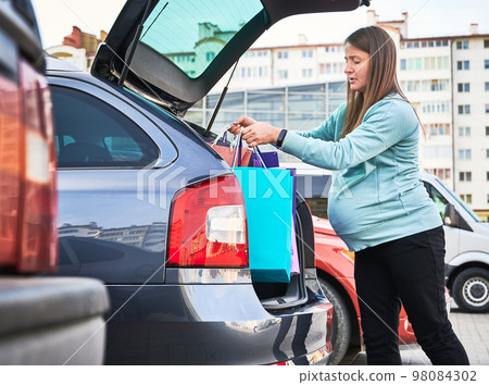 Side view of Caucasian woman with belly puting shopping bags in automobile trunk enjoying maternity and parenthood in awaiting mode. Pregnant female preparing for baby birth. 98084302