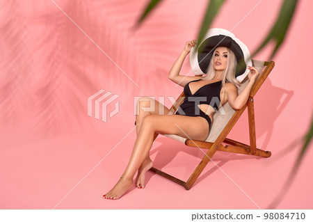 Young woman in black swimsuit relaxing on chaise longue, studio shot. Blurred palm tree leaves in foreground. 98084710