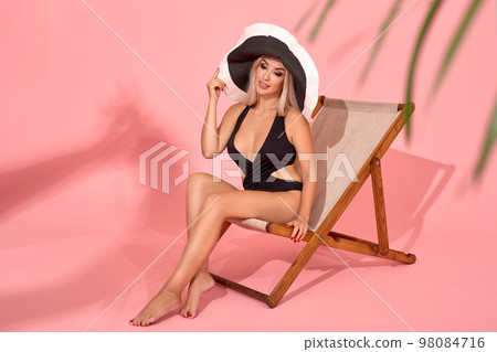 Young woman in black swimsuit relaxing on chaise longue, studio shot. Blurred palm tree leaves in foreground. 98084716