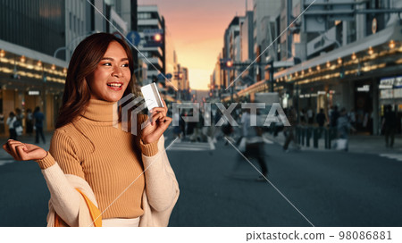 Elegant fashionable woman with credit card in hand standing against background of busy city street and evening sky 98086881