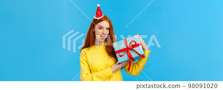 Check-out my gift. Sassy and daring good-looking redhead woman show-off, bragging with wrapped christmas present, looking camera happy and cheeky, wearing cute new year hat, blue background Check-out my gift. Sassy and daring good-looking redhead woman show-off, bragging with wrapped christmas present, looking camera happy and cheeky, wearing cute new year hat, blue background 98091020