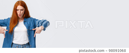 Wait a minute what is that. Frustrated and angry tensed redhead girlfriend asking wtf going on, dont understand, looking perplexed and mad down, pointing bottom copy space, stand white background 98091068