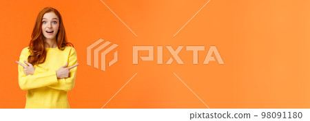 Amused dreamy and cute redhead girl asking advice in shopping mall, buying holiday presents, want valentines day gift, pointing sideways left and right, smiling upbeat, orange background 98091180